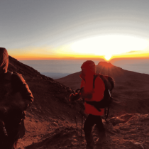 Two hikers in jackets and backpacks trek a rocky mountain trail at sunrise, bathed in golden light—perfectly capturing the spirit of a Kilimanjaro Expedition as they strive for new heights.